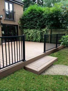 Grassy garden area led with a path to raised brown decking area and black railing at the back of a house.